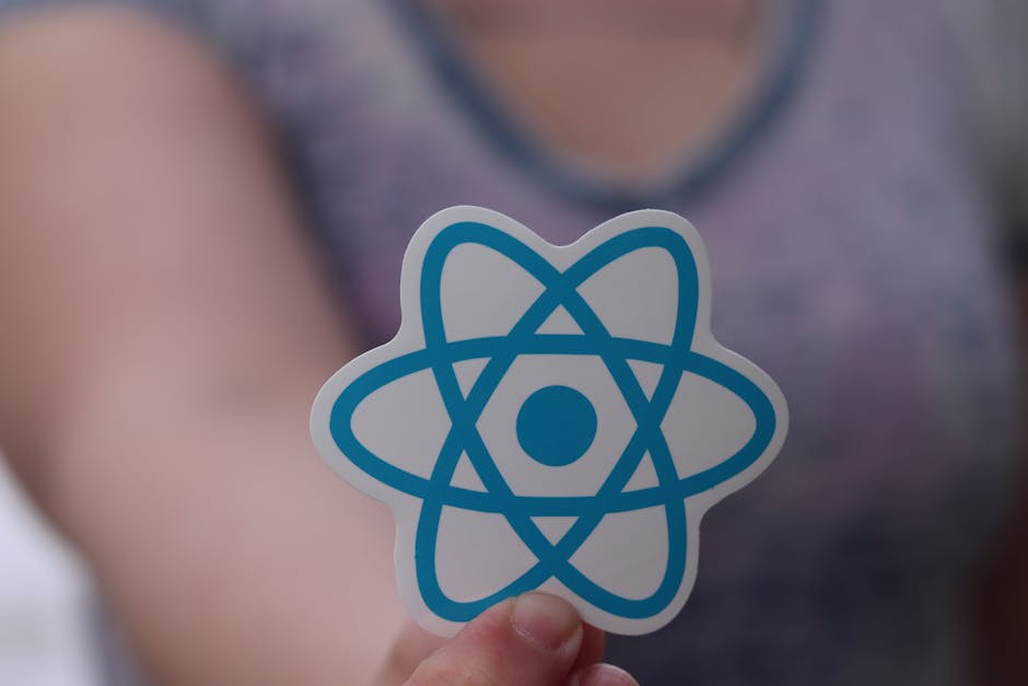 Close-up of a person's hand holding a small, printed sticker featuring the blue and white logo of an atom, with a blurred background showing the person's arm and grey clothing. The setting appears ind